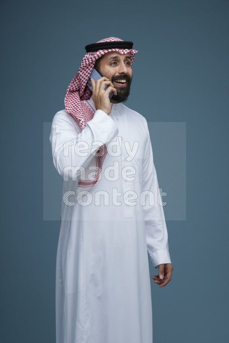 A man having a Call in a blue background wearing Saudi Thob with Shomag