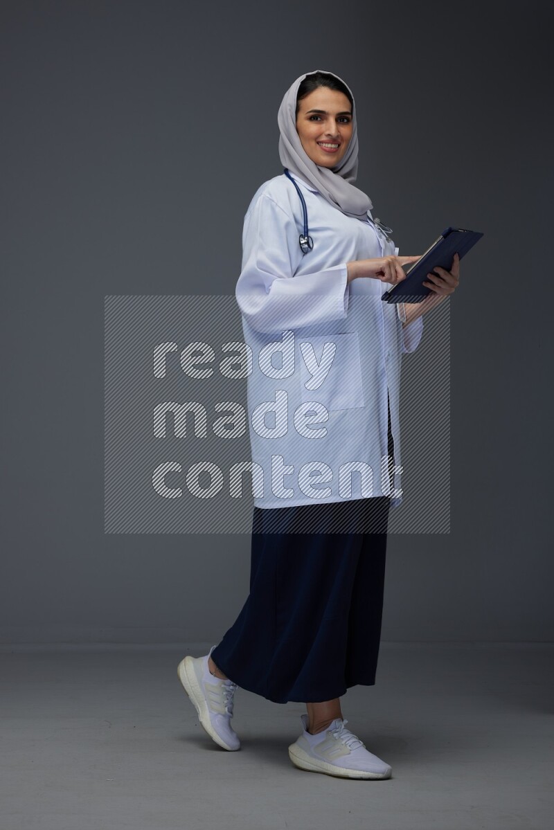 A Saudi doctor wearing a light gray head scarf standing and crossing her hands eye level on a grey background
