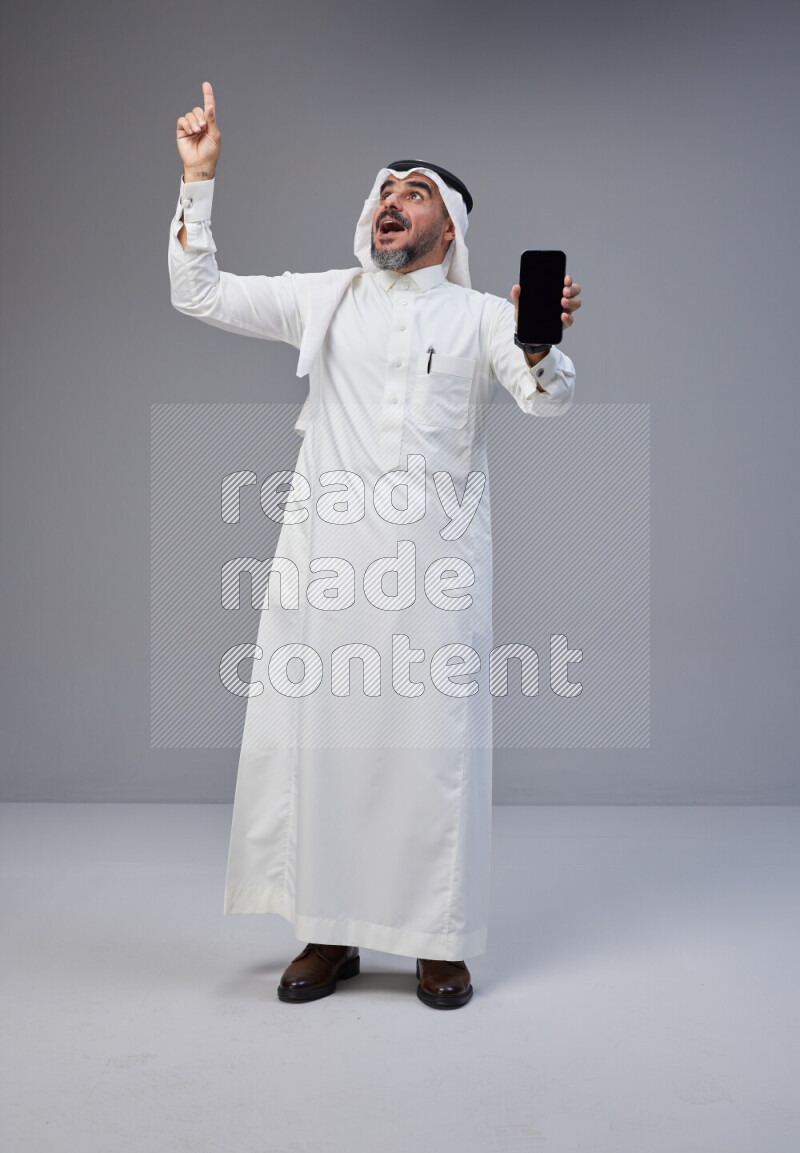 Saudi man Wearing Thob and white Shomag standing showing phone to camera on Gray background