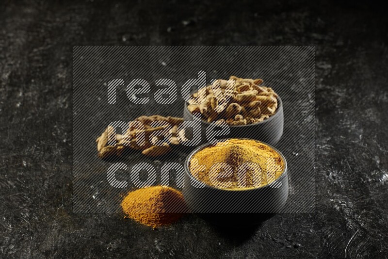 2 pottery black bowls, one full of turmeric powder and the other full of dried turmeric whole fingers on a textured black flooring