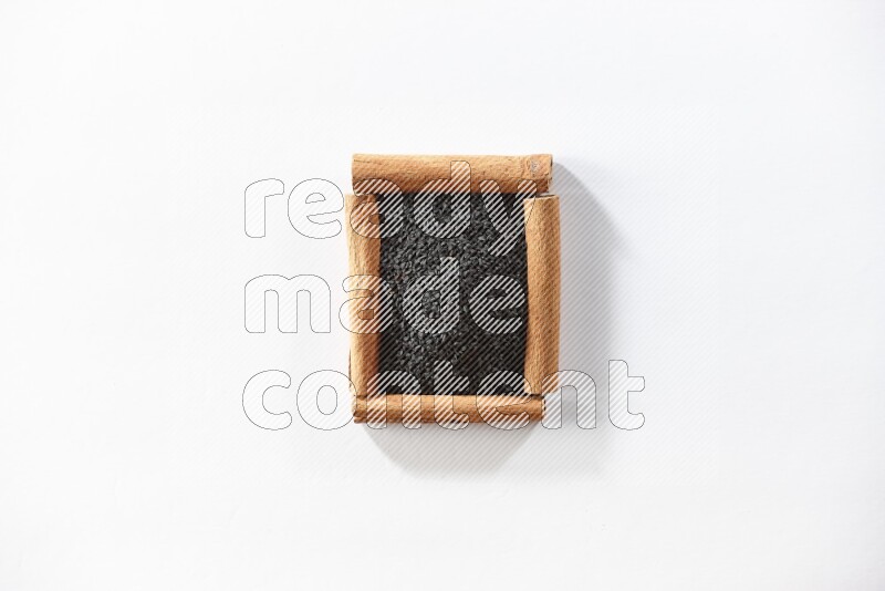 أربعة من أعواد القرفة مكونة شكل مربع بداخلها حبة البركة علي خلفية بيضاء