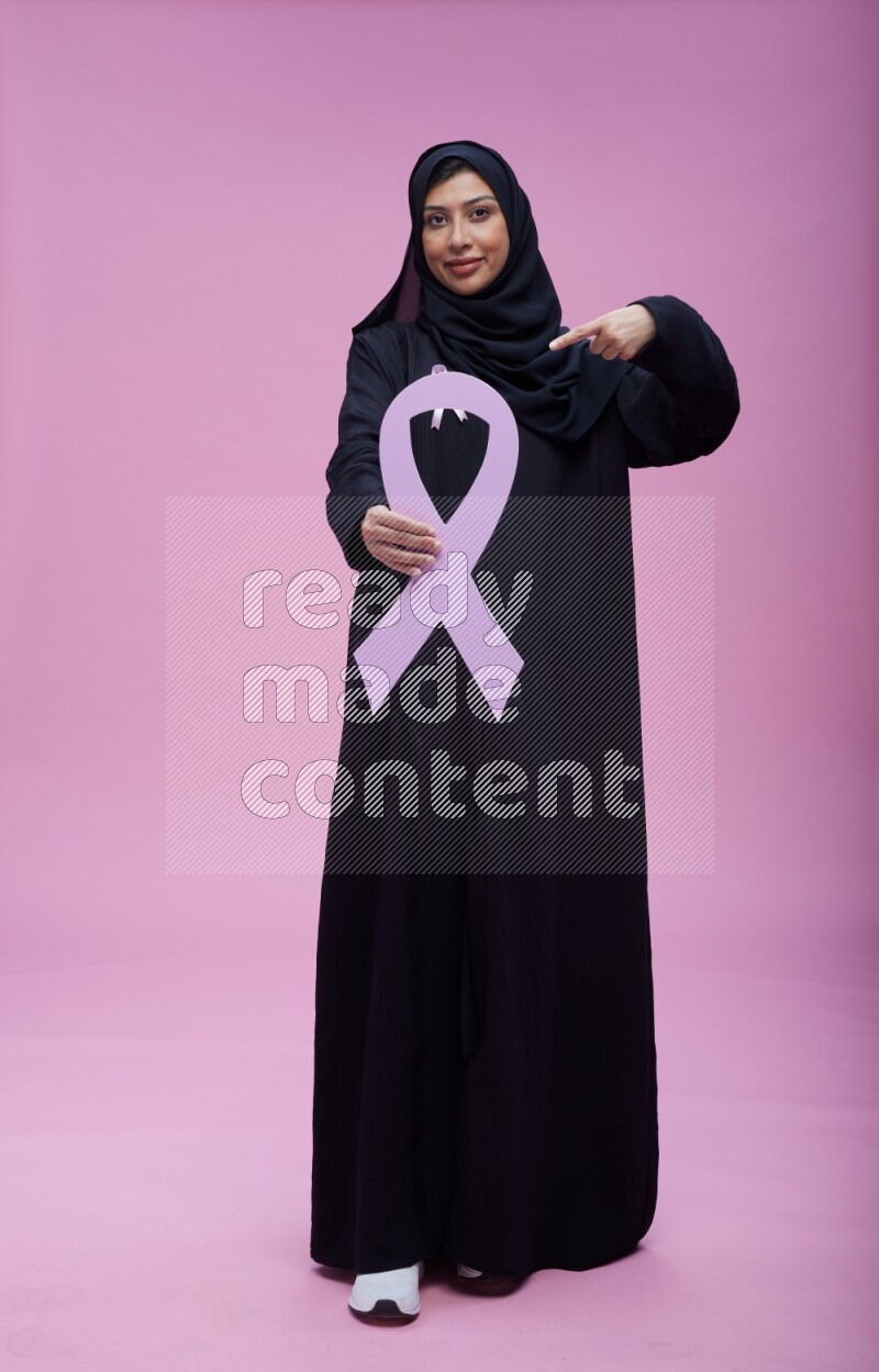 Saudi woman wearing pink ribbon on abaya standing holding awareness ribbon on plain pink background