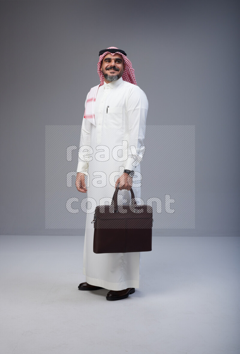 Saudi man Wearing Thob and red Shomag standing holding bag on Gray background