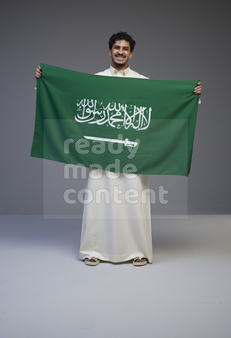 A saudi man standing wearing thob holding big saudi flag on gray background