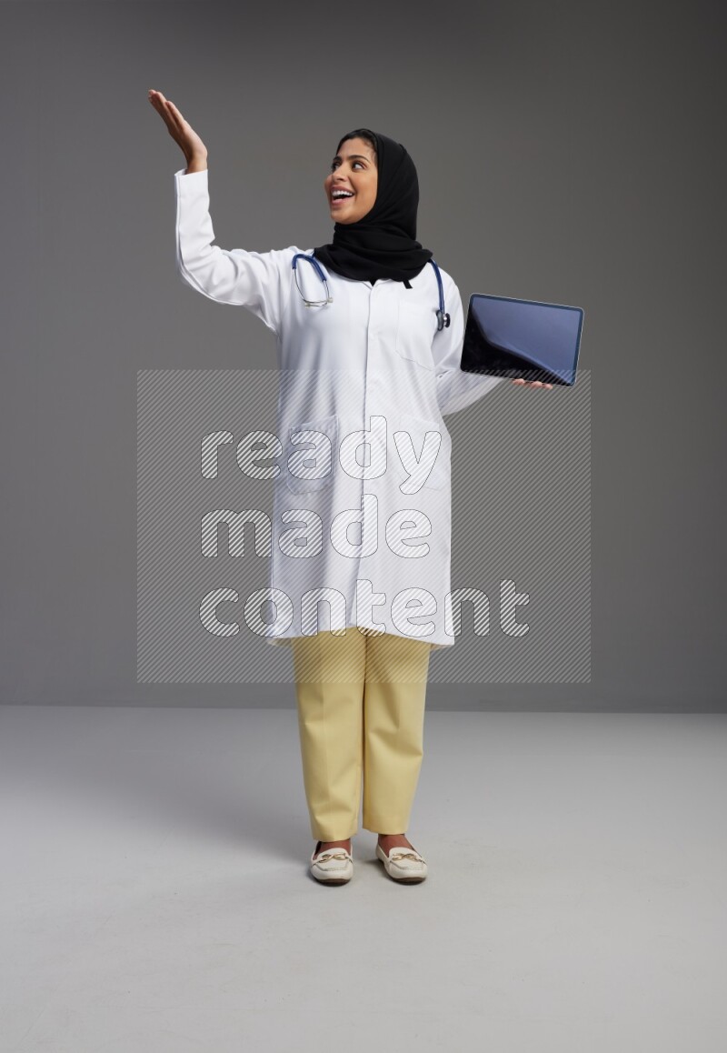 Saudi woman wearing lab coat with stethoscope standing showing tablet to camera with sign in the back on Gray background