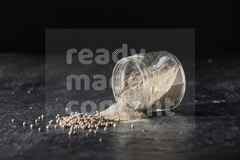 A flipped glass jar full of white pepper powder with spilled powder and pepper beads on textured black flooring