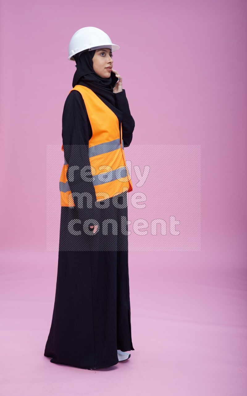 Saudi woman wearing Abaya with engineer vest and helmet standing talking on phone on pink background