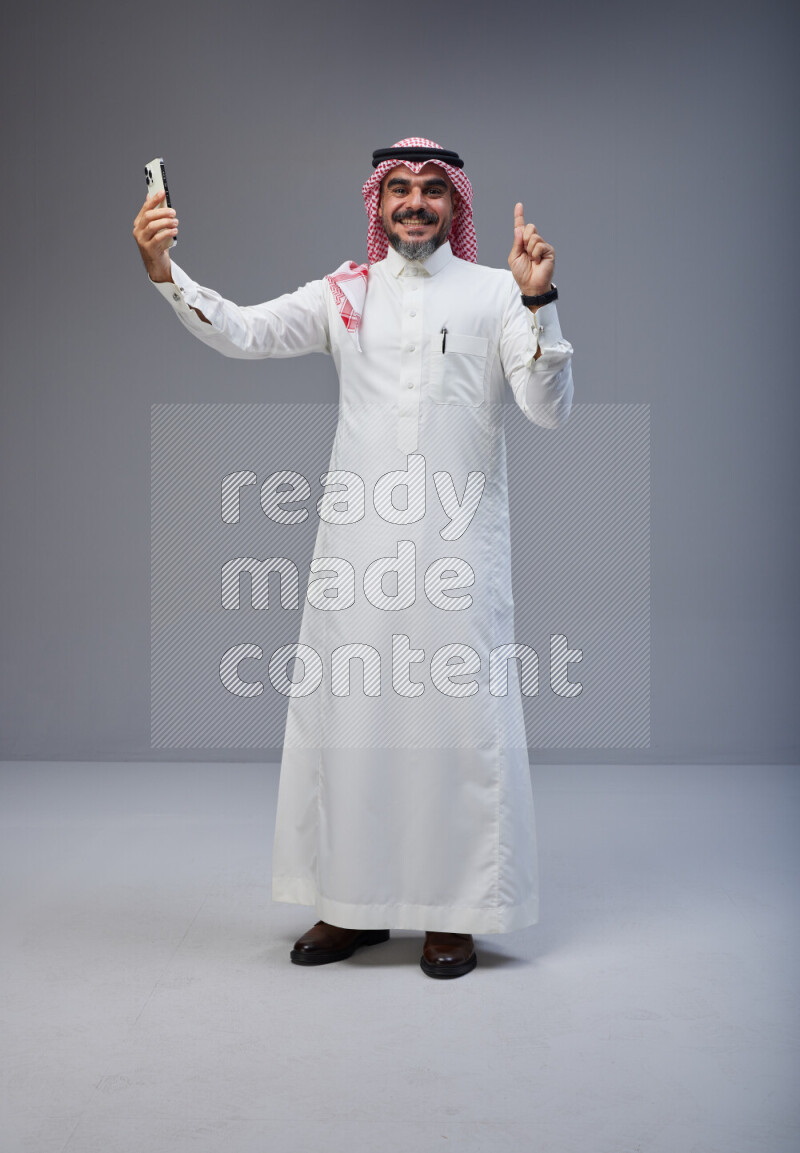 Saudi man Wearing Thob and red Shomag standing taking selfie on Gray background