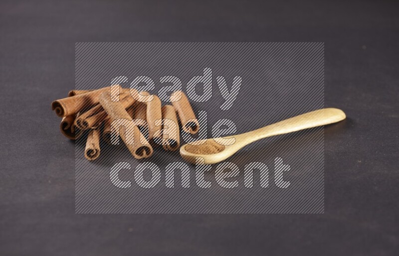 Cinnamon sticks stacked beside a wooden spoon full of cinnamon powder on black background