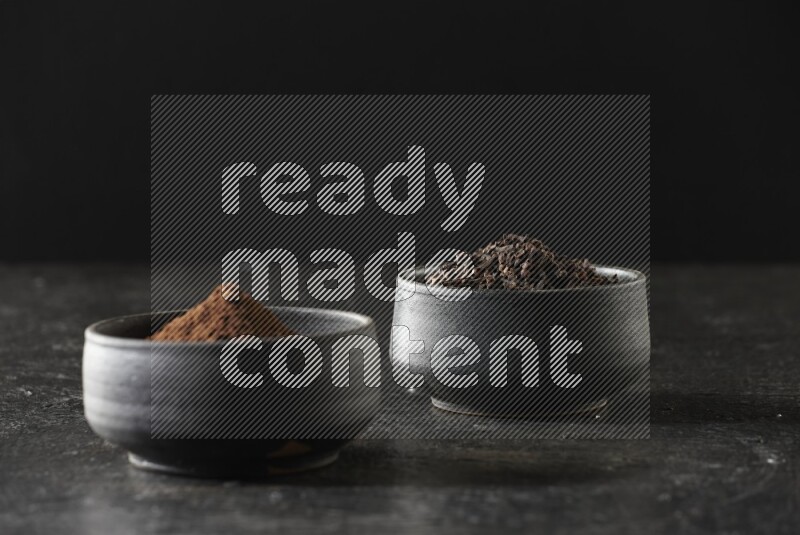 2 Black pottery bowls full of cloves and the other full of cloves powder on textured black flooring