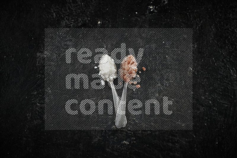 ملعقتان معدنيتان ممتلئتان بالملح الأبيض وملح الهيمالايا الوردي على خلفية سوداء