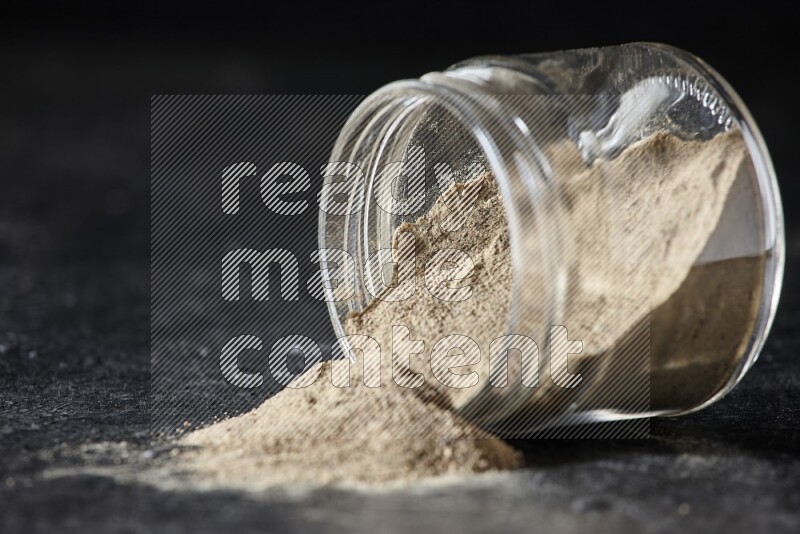 A flipped glass jar full of white pepper powder with spilled powder on textured black flooring