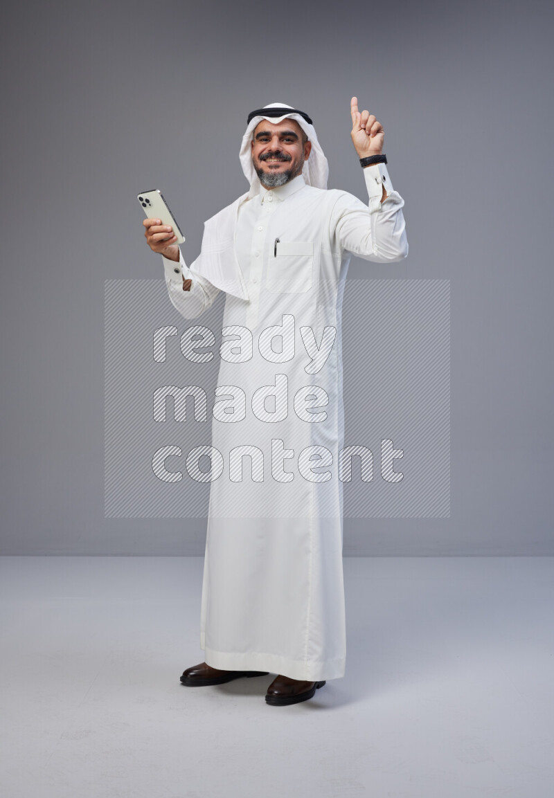 Saudi man Wearing Thob and white Shomag standing texting on phone on Gray background