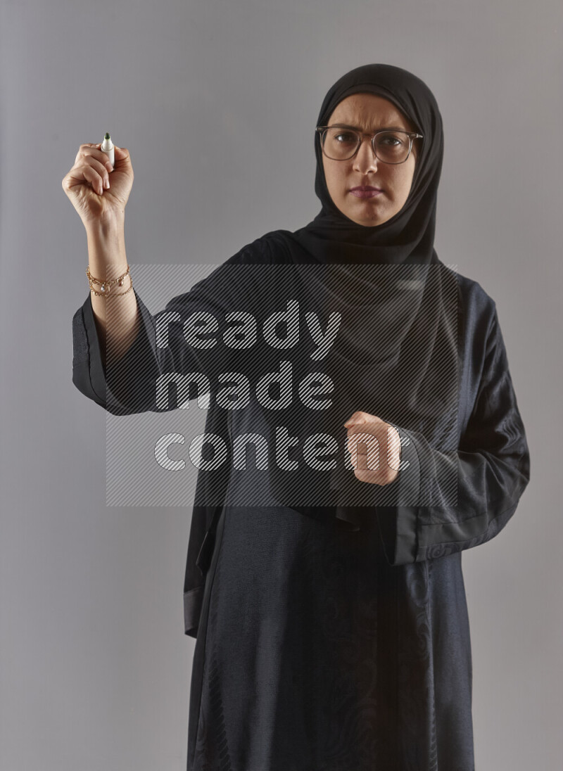 Woman in black abaya, hijab and glasses holding a marker pen to write on a transparent board with different reactions and expressions on grey background