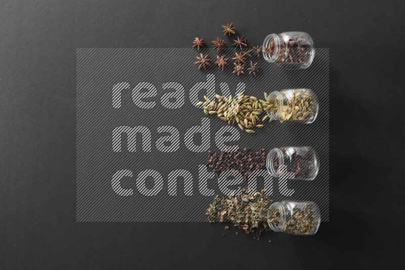 4 glass spice jars full of cloves, cardamom, basil and star anise on black flooring