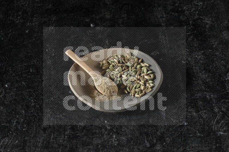 A plate filled with cardamom seeds and a wooden spoon full of cardamom powder on a textured black flooring