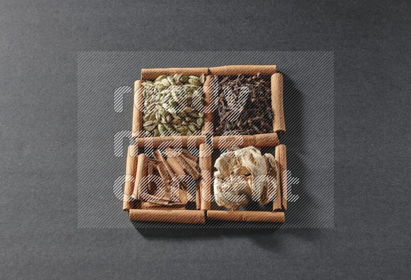 4 مربعات من عيدان القرفة ممتلئة بقطع القرفة وحبوب الهيل والقرنفل والزنجبيل المجفف على أرضية سوداء