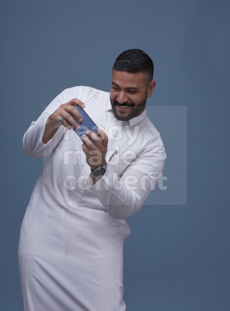 A man Playing a Game on his Smartphone  on Blue Background wearing Saudi Thob
