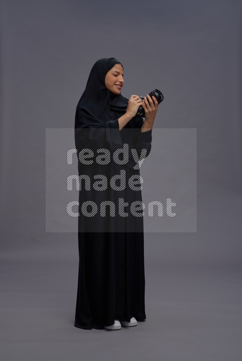 Saudi woman wearing Abaya standing holding Camera on gray background