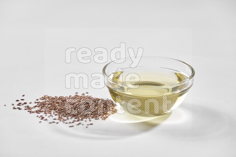 A glass bowl full of flaxseeds oil and flaxseeds beside it on a white flooring