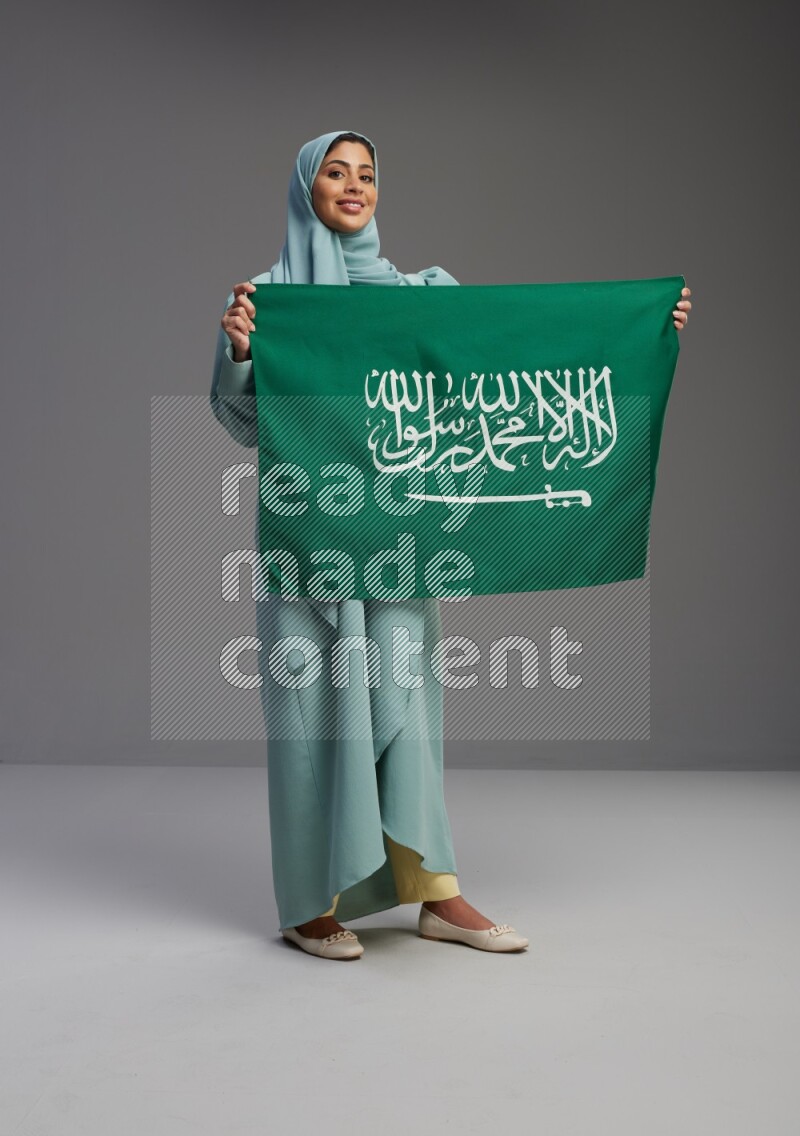 Saudi Woman wearing Abaya standing holding Saudi flag on Gray background