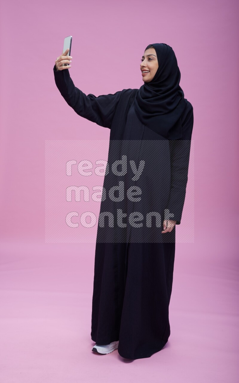 Saudi woman wearing Abaya standing taking selfie on pink background