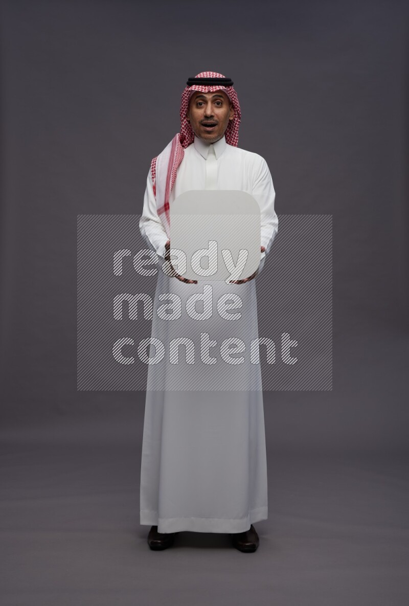 Saudi man wearing thob and shomag standing holding social media sign on gray background