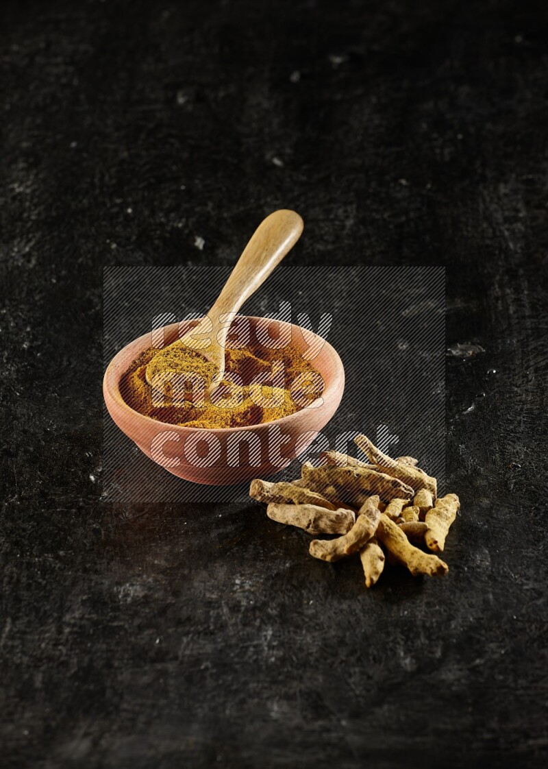 A wooden bowl and a wooden spoon full of turmeric powder with dried turmeric fingers on textured black flooring