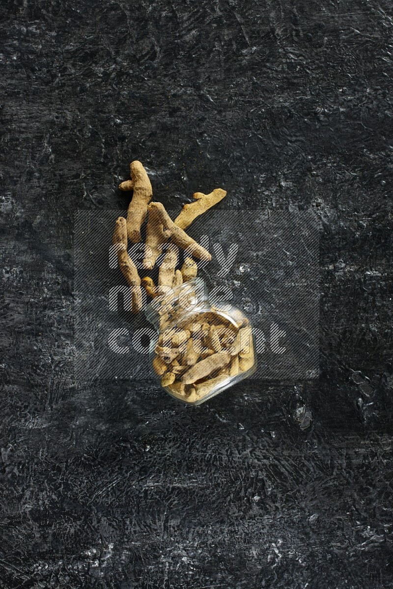 وعاء زجاجي للتوابل مقلوب وممتلئ بأصابع الكركم المجففة وبعض أصابع الكركم خارج الوعاء على خلفية سوداء