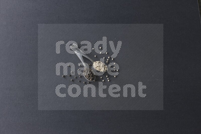 2 metal spoons full of black and white pepper beads with spreaded beads on black flooring