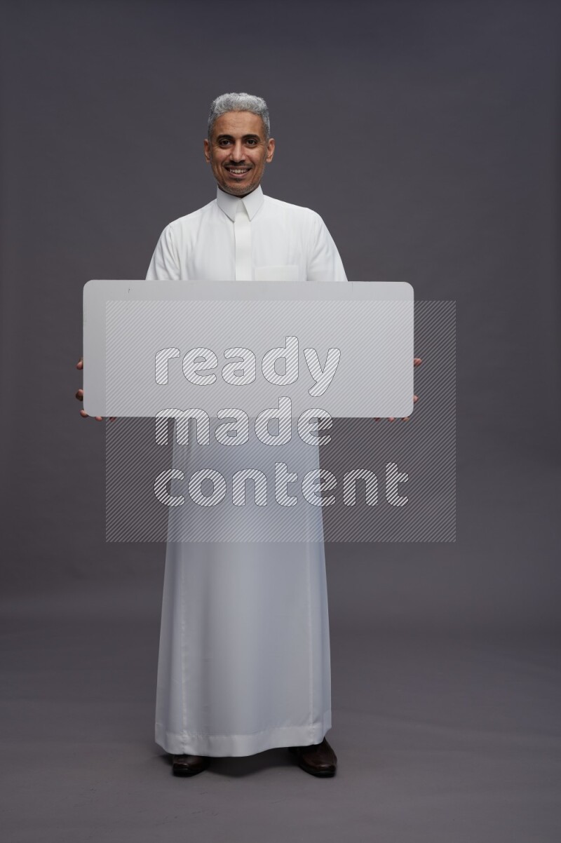 Saudi man wearing thob standing holding board on gray background