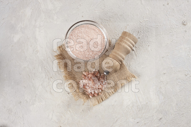A glass bowl full of pink himalayan salt with a wooden grinder on a burlap fabric all on white background