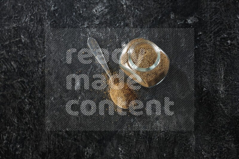 وعاء زجاجي للتوابل وملعقة معدنية ممتلئين ببودرة الكمون على خلفية سوداء