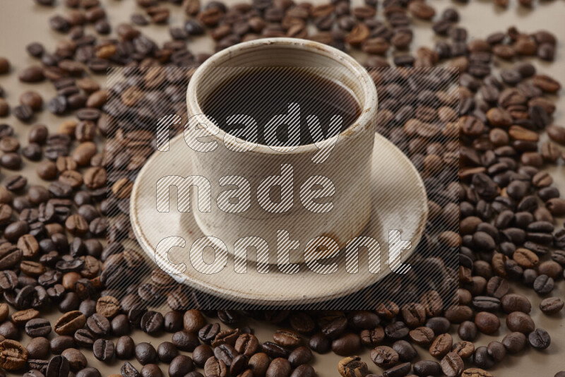 A beige pottery cup of coffee surrounded by roasted coffee beans on beige background