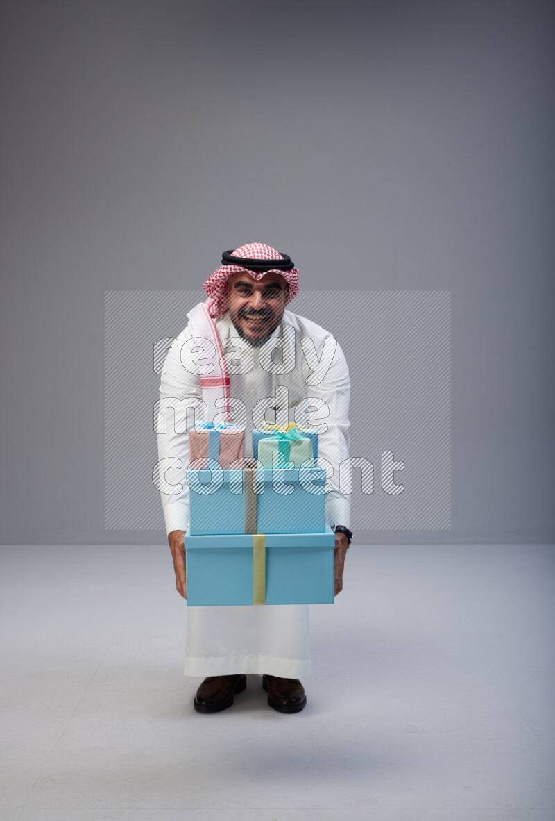Saudi man Wearing Thob and red Shomag standing holding gift box on Gray background