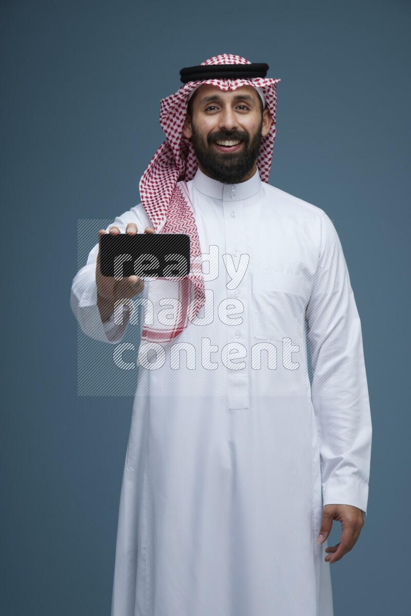 A man showing his phone Screen in a blue background wearing Saudi Thob with Shomag