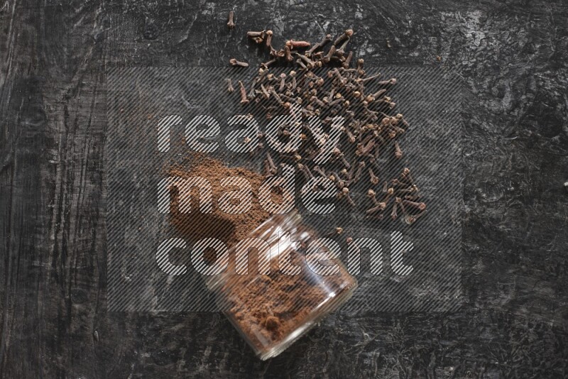 A flipped glass jar full of cloves powder with cloves spread on a textured black flooring