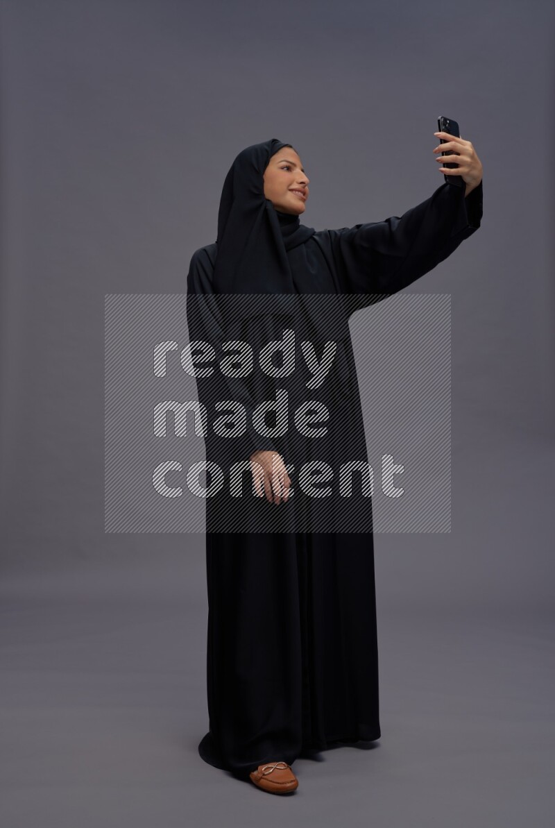 Saudi woman wearing Abaya standing taking selfie on gray background