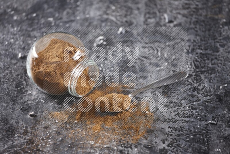 Herbal glass jar full of cinnamon powder flipped and a metal spoon on textured black background