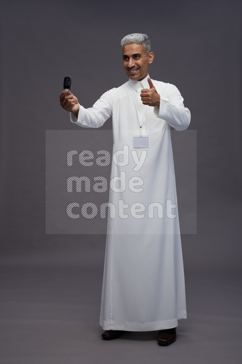 Saudi man wearing thob with neck strap employee badge standing holding key car on gray background