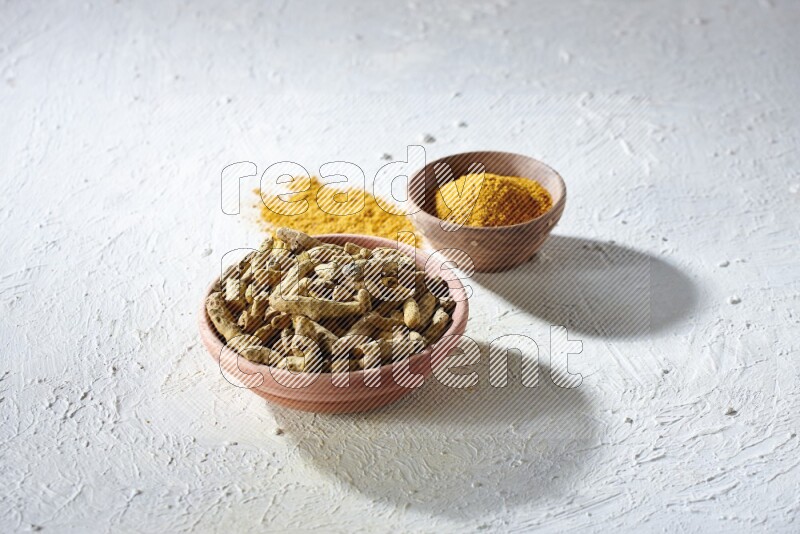 2 wooden bowls, one full of turmeric powder and the other full of dried turmeric whole fingers on a textured white flooring