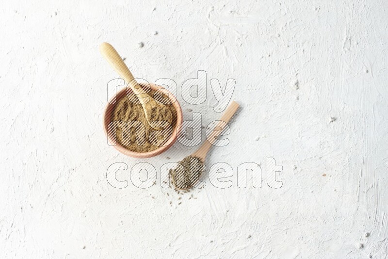 A wooden bowl and 2 wooden spoons full of cumin powder and cumin seeds on textured white flooring