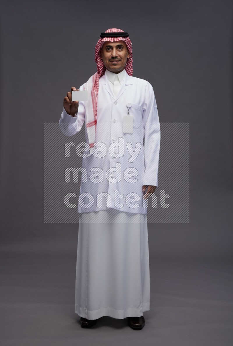 Saudi man wearing thob with lab coat and shomag with pocket employee badge standing holding ATM card on gray background