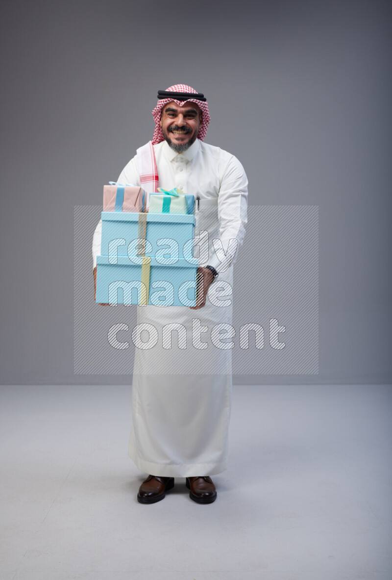 Saudi man Wearing Thob and red Shomag standing holding gift box on Gray background