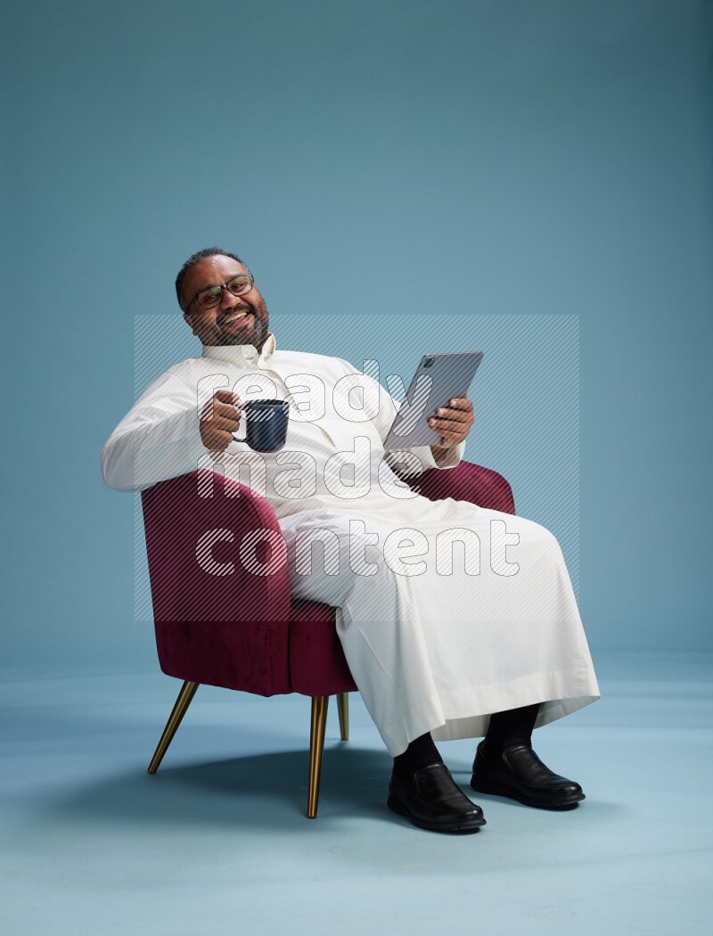 Saudi Man without shimag sitting on chair working on tablet on blue background