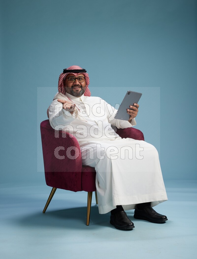 Saudi Man with shimag sitting on chair working on tablet on blue background