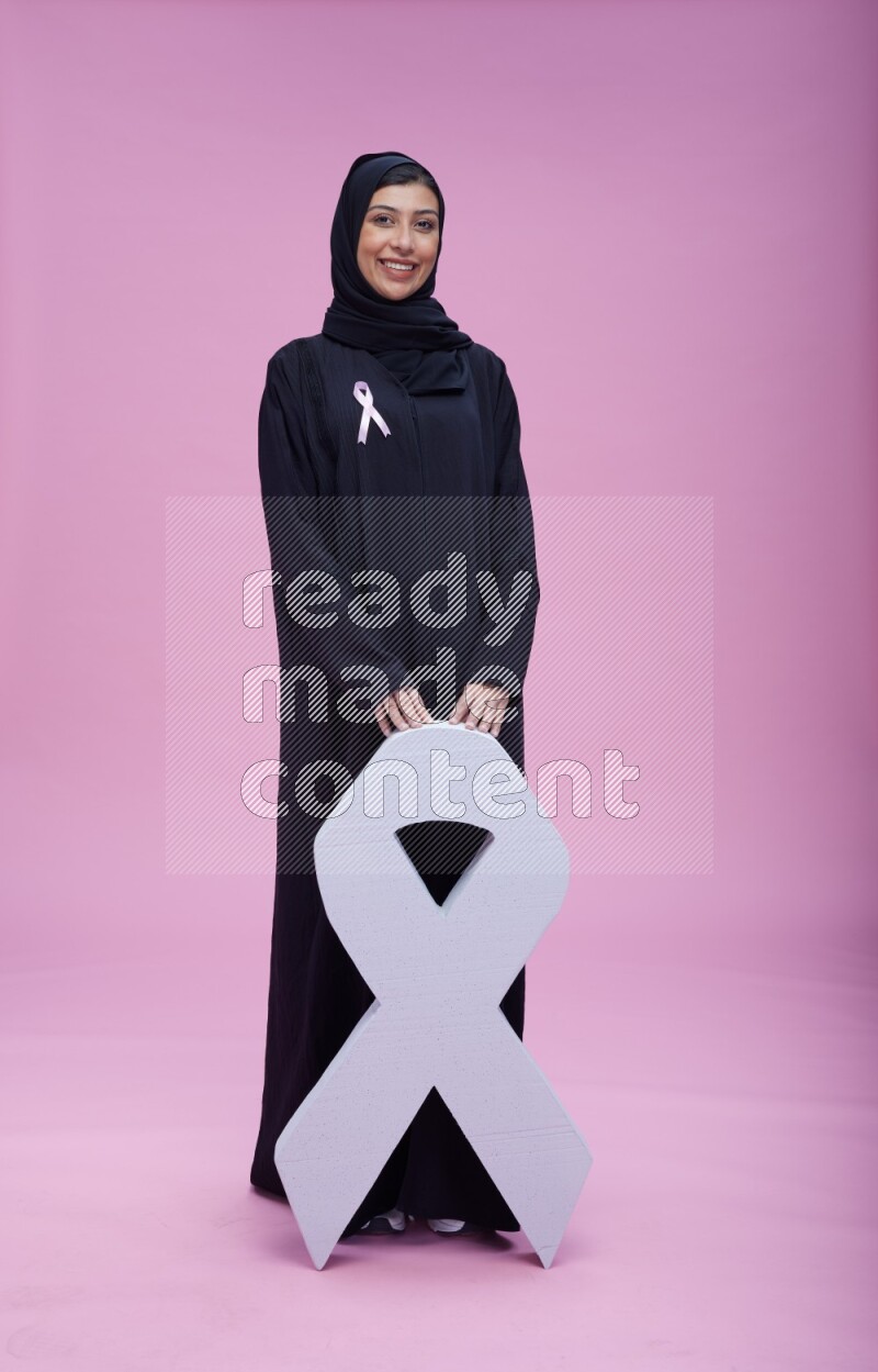 Saudi woman wearing pink ribbon on abaya standing holding awareness ribbon on plain pink background