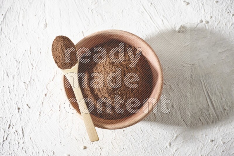 A wooden bowl and a wooden spoon full of cloves powder on a textured white flooring