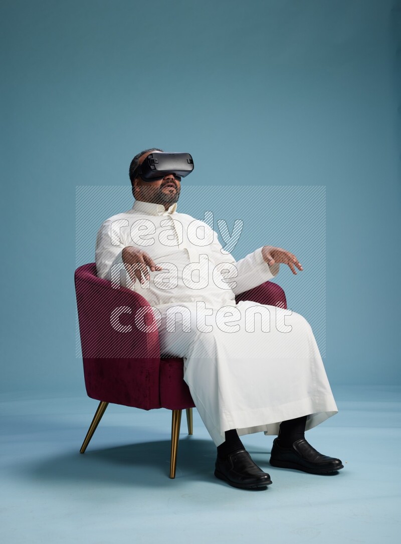 Saudi Man without shimag sitting on chair  gaming with VR on blue background