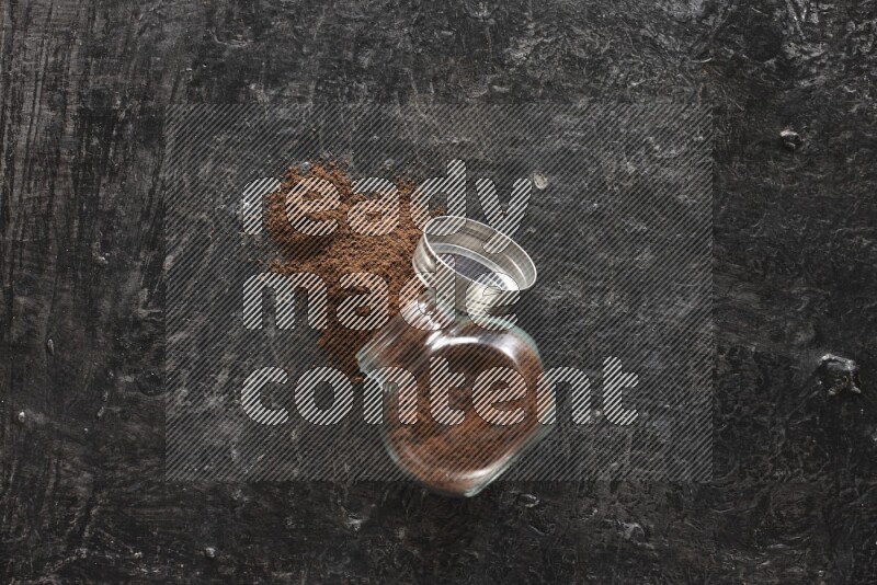 A flipped glass spice jar full of cloves powder and powder came out of it on textured black flooring